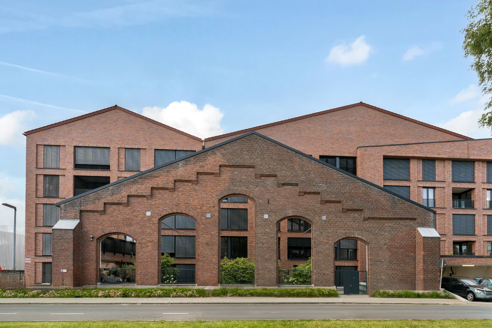 Modernes Wohngebäude aus rotem Backstein mit großen Fenstern, davor eine Straße.