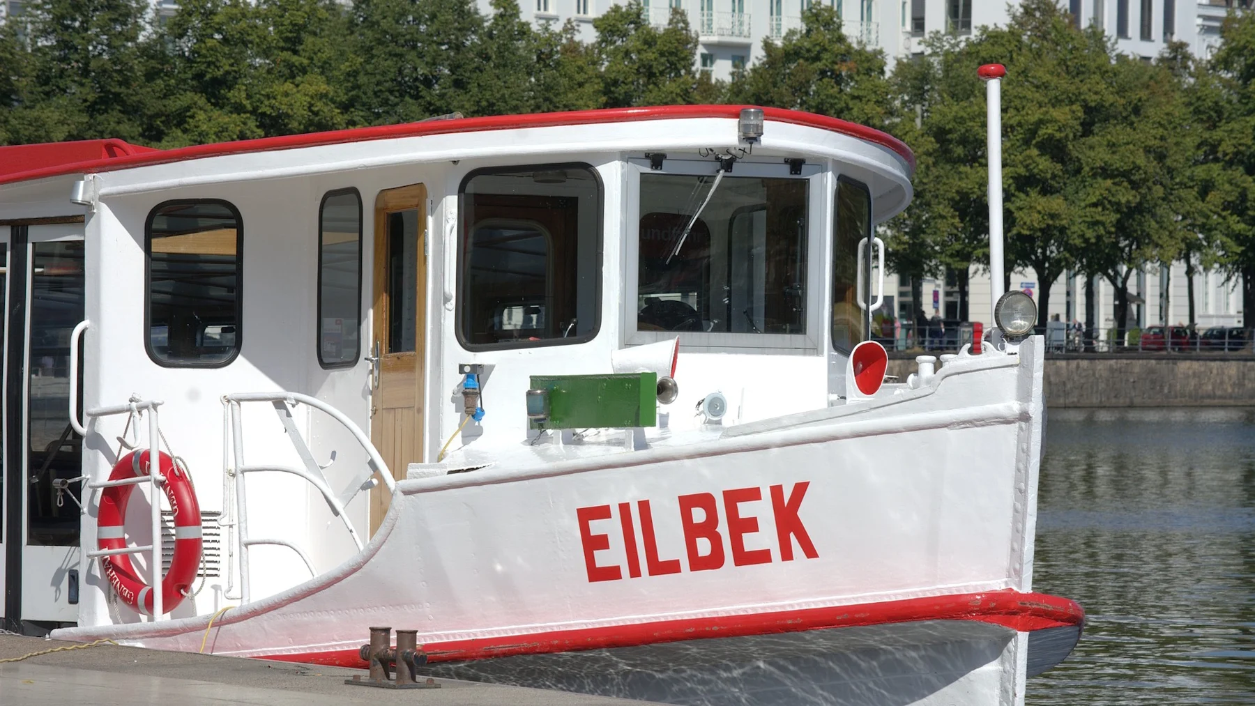 Weißes Boot mit rotem Dach und Schriftzug 'EILBEK' am Ufer, im Hintergrund Bäume und Gebäude.