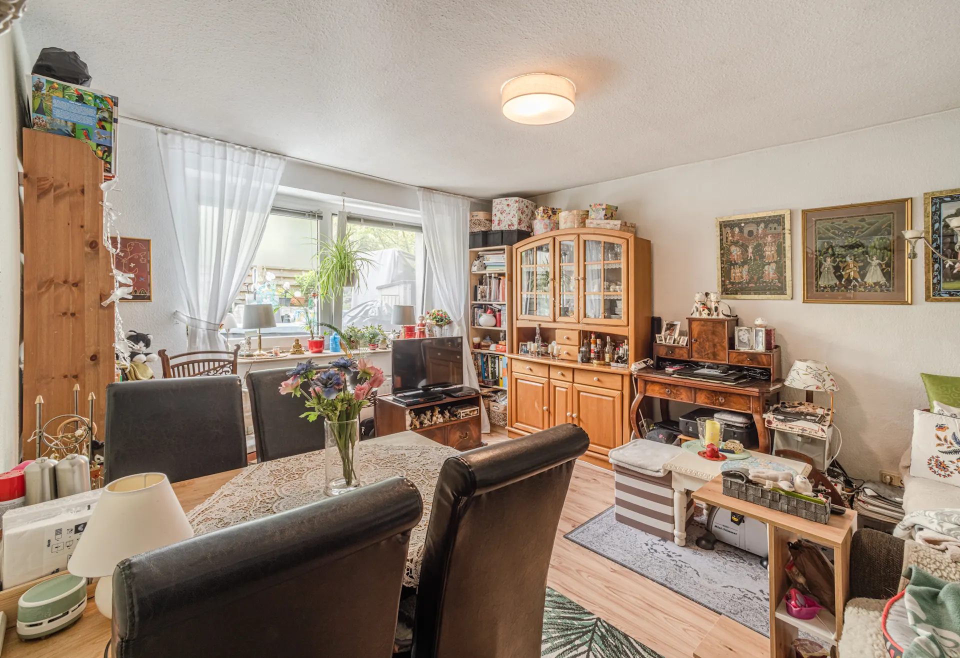 Wohnzimmer mit Esstisch, Blumen, Schränken, Sofa, Fernseher und Fenstern mit weißen Vorhängen.