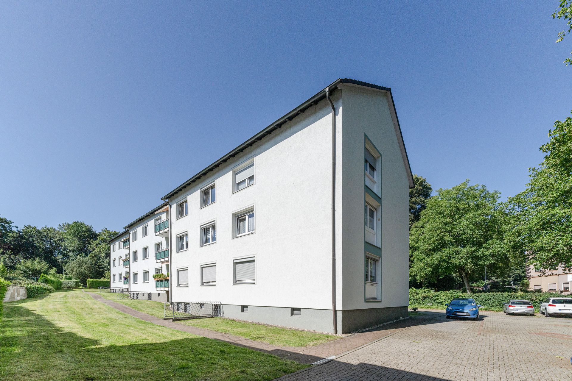 Mehrfamilienhaus mit weißen Fassaden, Grünfläche und geparkten Autos bei sonnigem Wetter.