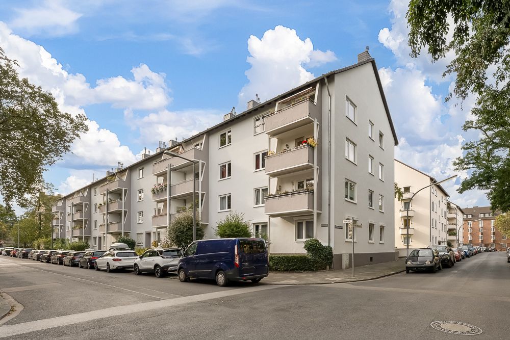 Mehrstöckiges Wohnhaus mit Balkonen an einer Straßenecke, geparkte Autos davor.