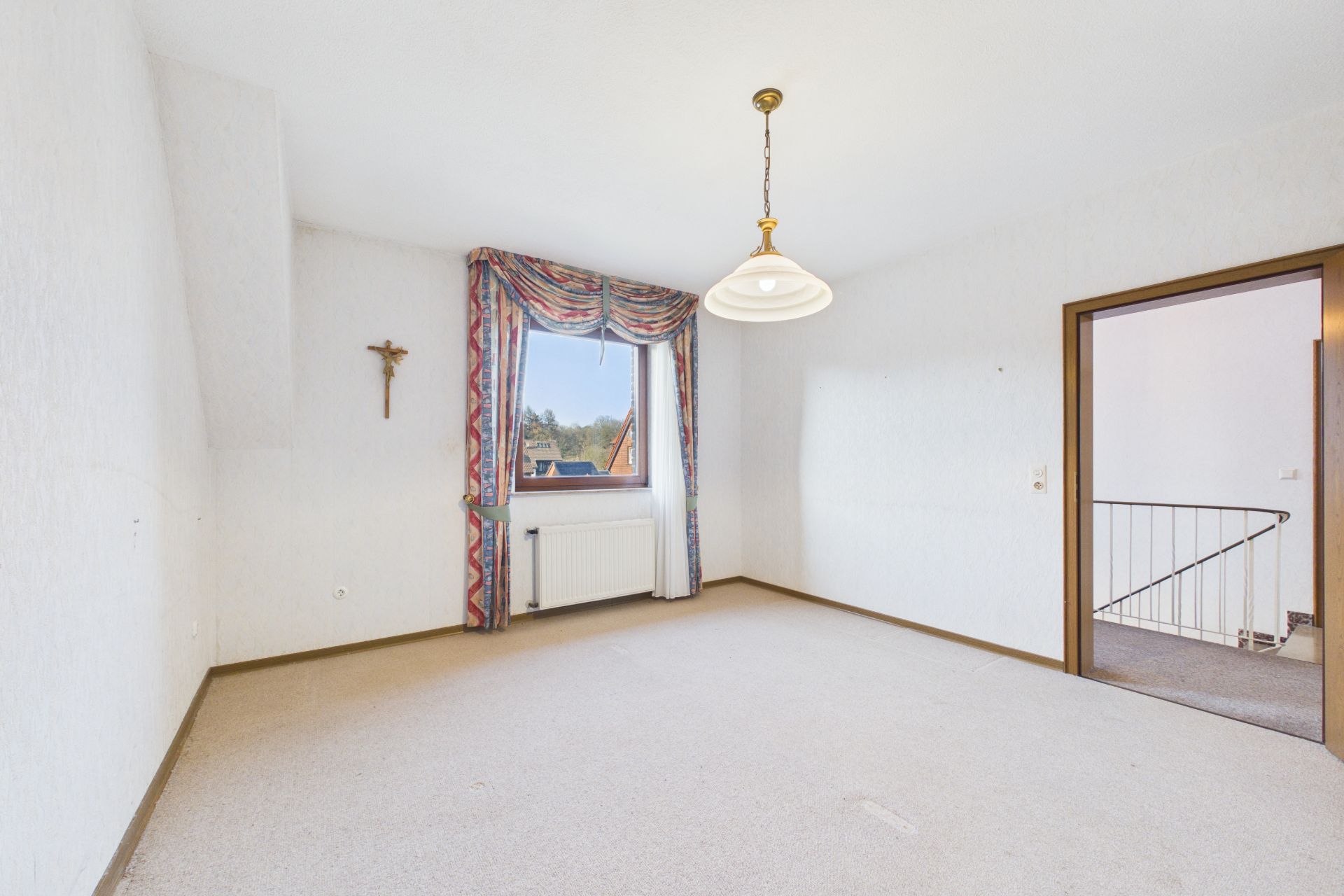 Heller, leerer Raum mit Teppichboden, Fenster mit Vorhang, Kreuz an der Wand und Deckenlampe.
