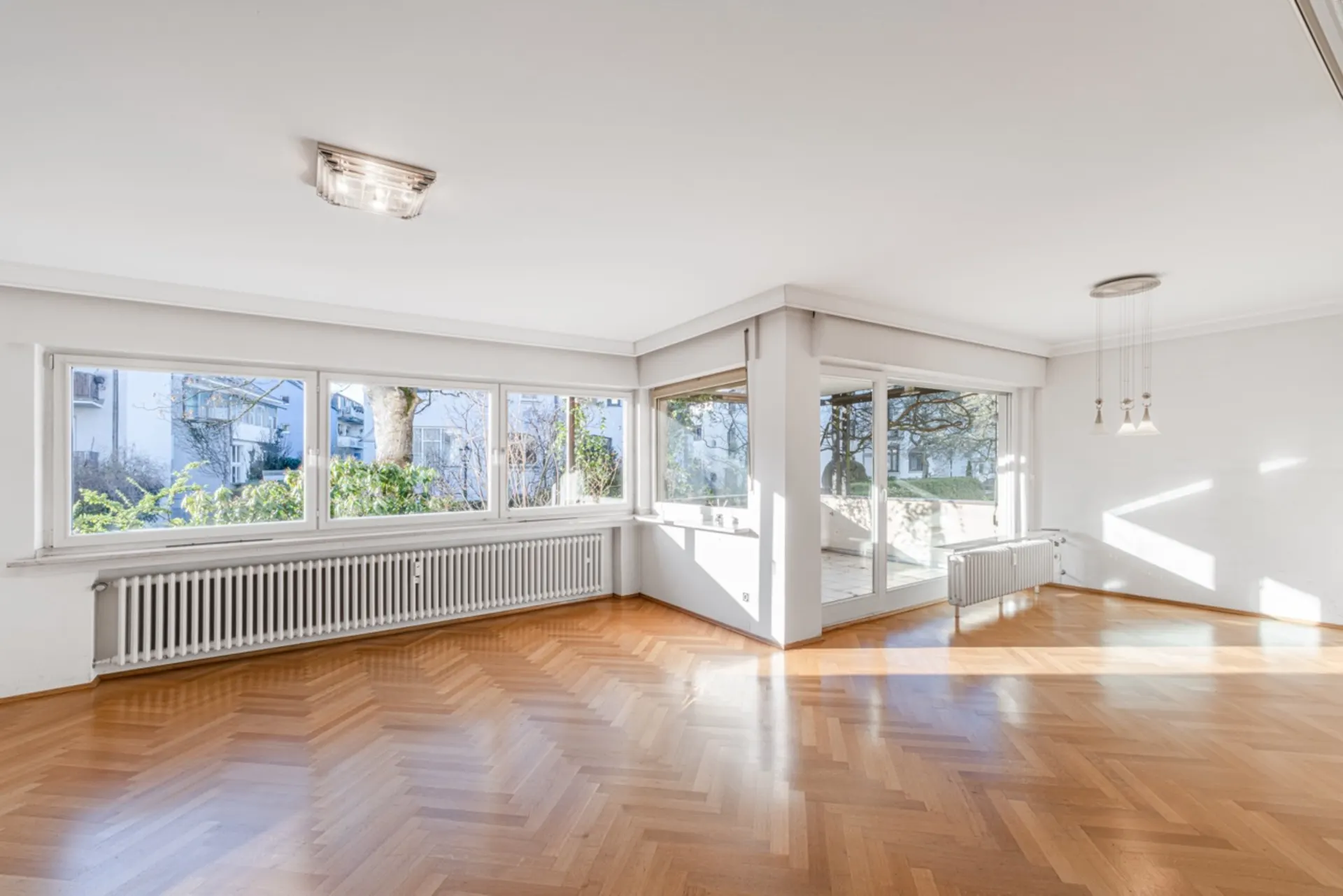 Heller, leerer Raum mit großen Fenstern, Parkettboden und Blick auf Garten.