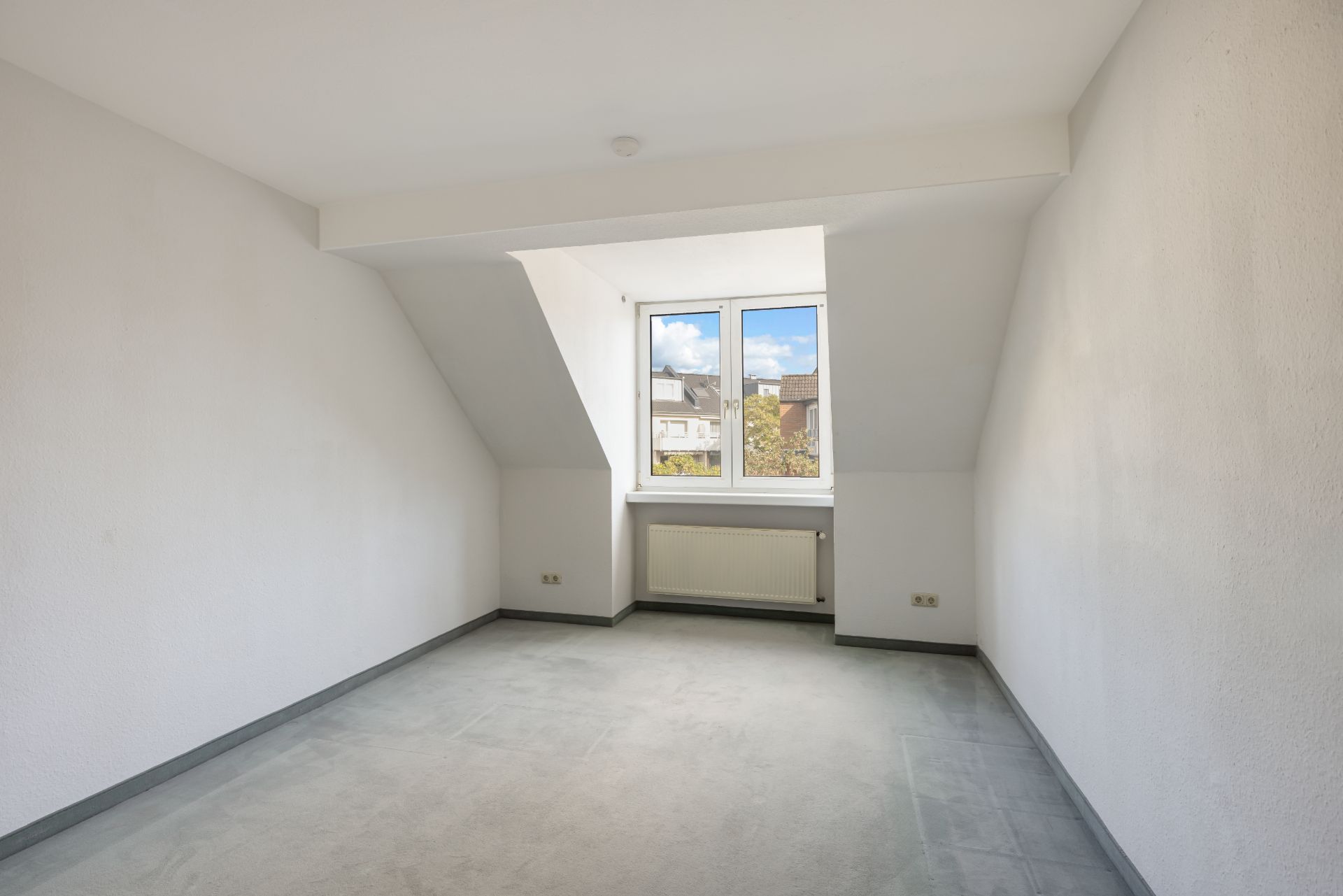 Leeres Zimmer mit Fenster, grauem Boden und weißen Wänden, unter dem Fenster befindet sich ein Heizkörper.