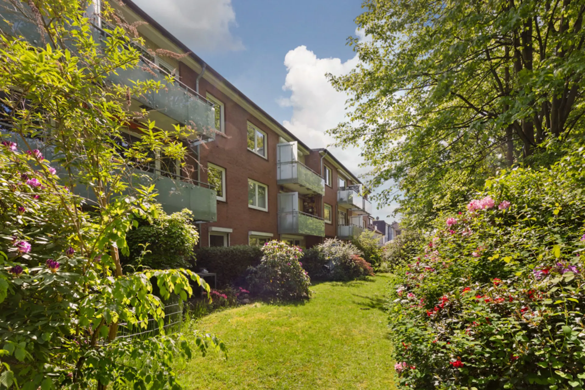 Mehrfamilienhaus mit Balkonen und gepflegtem Gartenbereich, umgeben von Bäumen und Blumen.