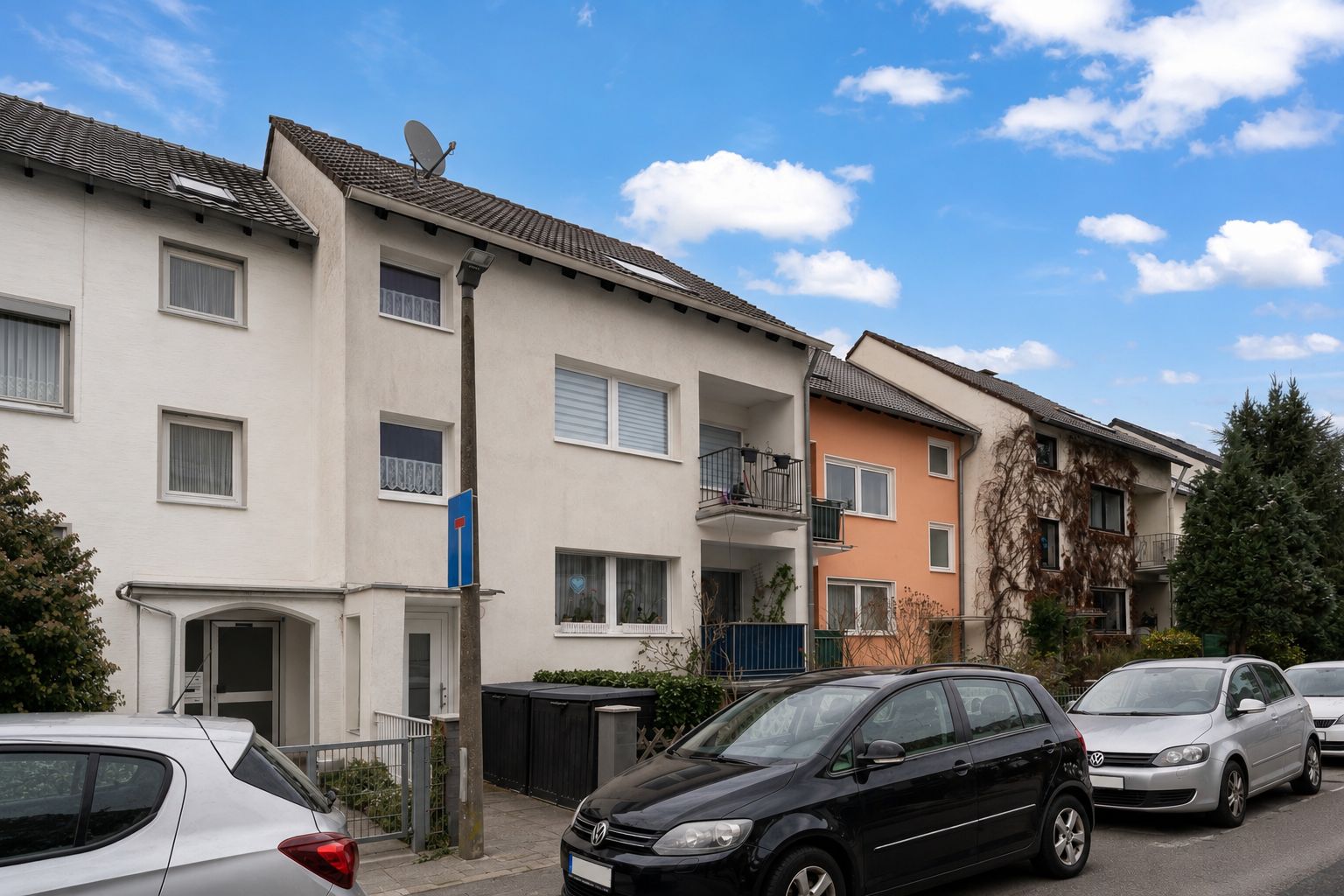 Mehrfamilienhaus mit Balkon und geparkten Autos an der Straße bei Tageslicht.