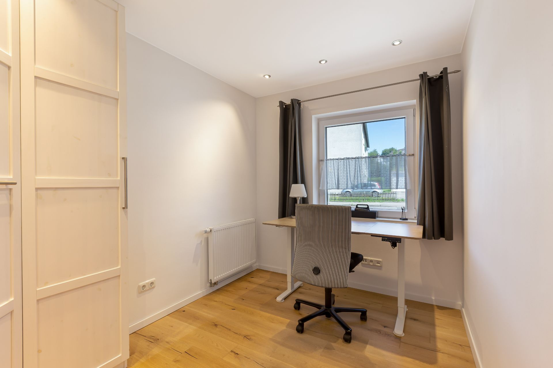 Kleines Arbeitszimmer mit Schreibtisch, Bürostuhl, Lampe und Fenster mit Vorhängen.