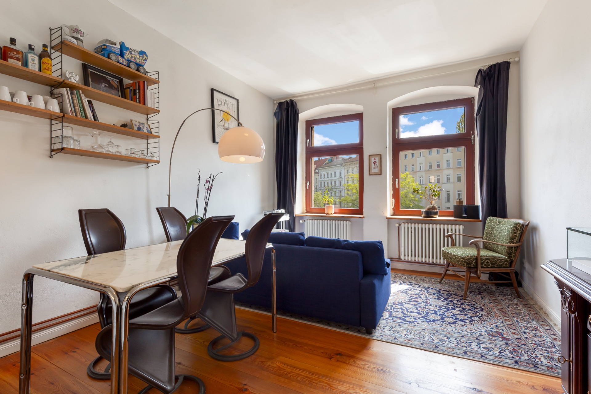 Helles Wohnzimmer mit Tisch, vier Stühlen, blauem Sofa, Teppich und drei großen Fenstern.