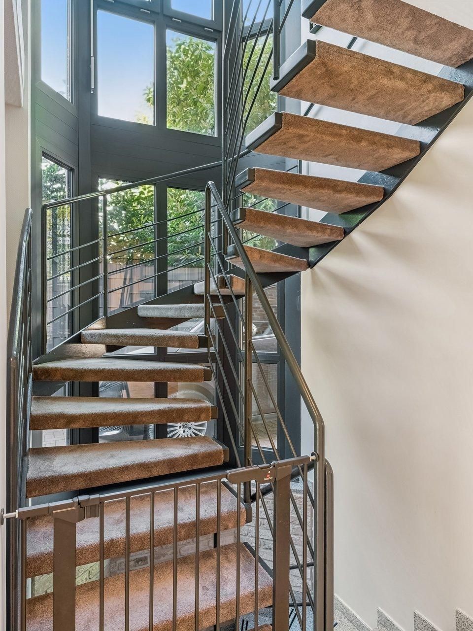 Moderne Wendeltreppe mit Metallgeländer und Teppichstufen vor großen Fenstern.