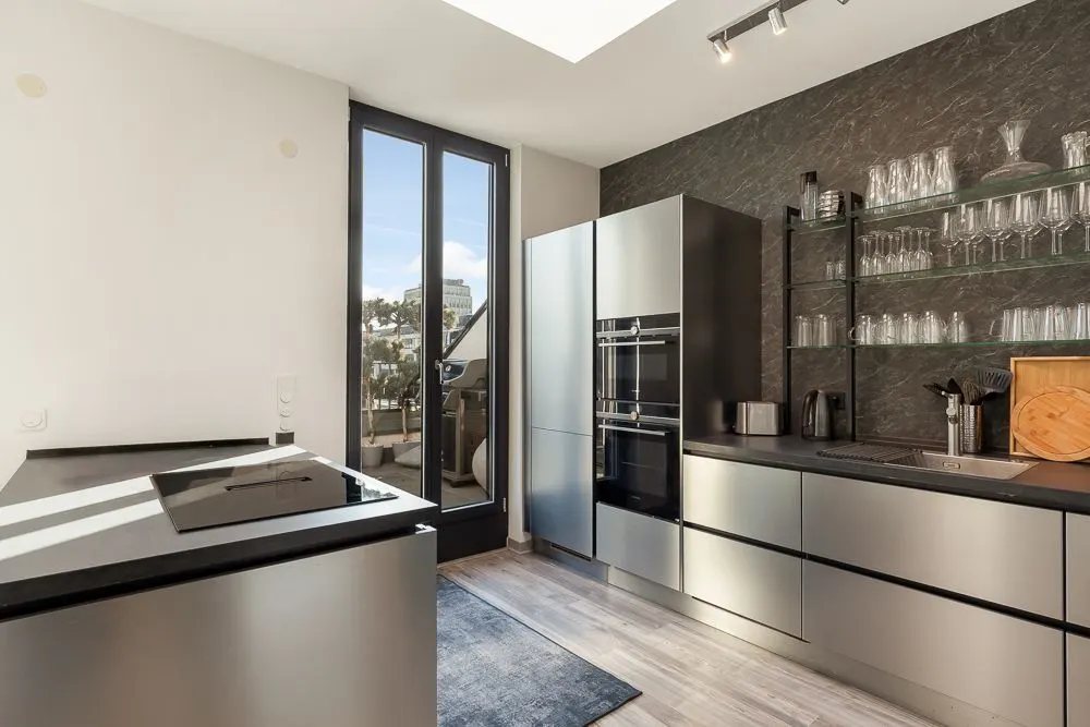 Moderne Küche mit Edelstahlgeräten, offenem Regal mit Gläsern und großem Fenster mit Zugang zum Balkon.