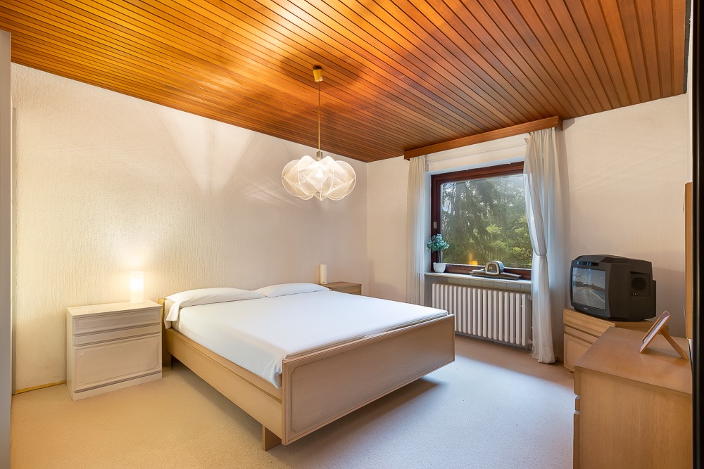 Schlafzimmer mit Doppelbett, Nachttischen, Holzdecke, Fenster und Fernseher auf Kommode.