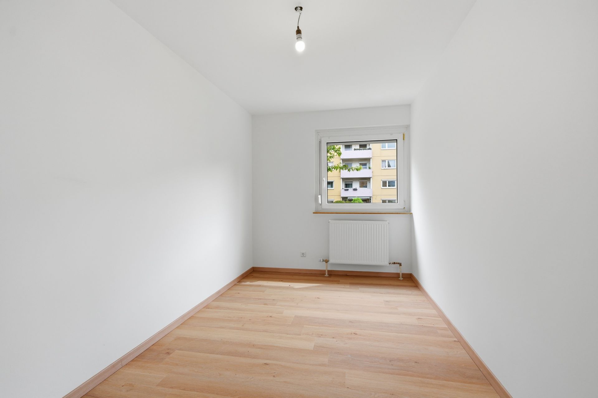 Leeres Zimmer mit Holzfußboden, weißer Wand, Fenster und Heizkörper.