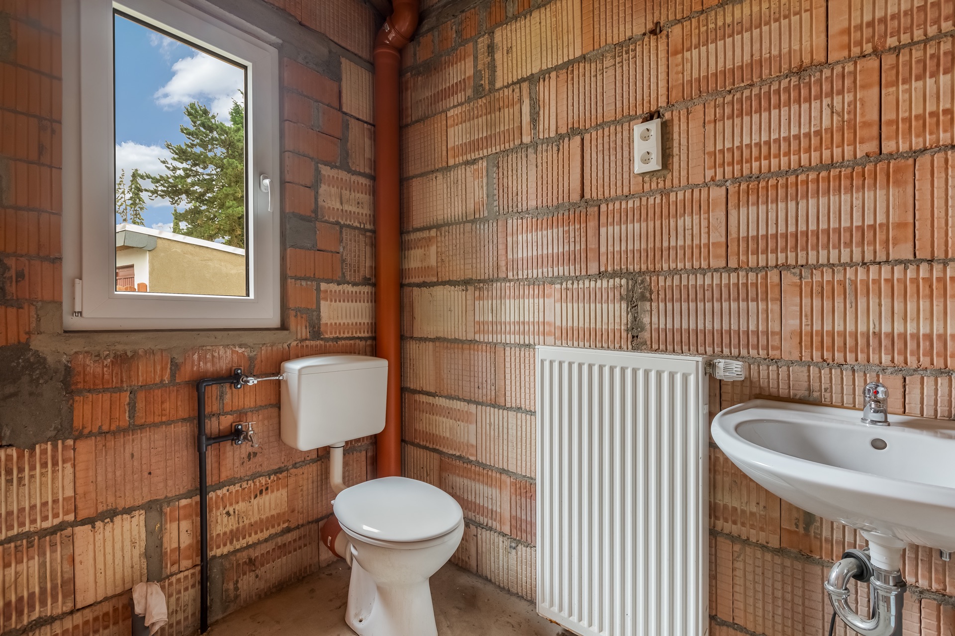 Toilette mit Spülkasten, Waschbecken und Heizung an unverputzter Ziegelwand.