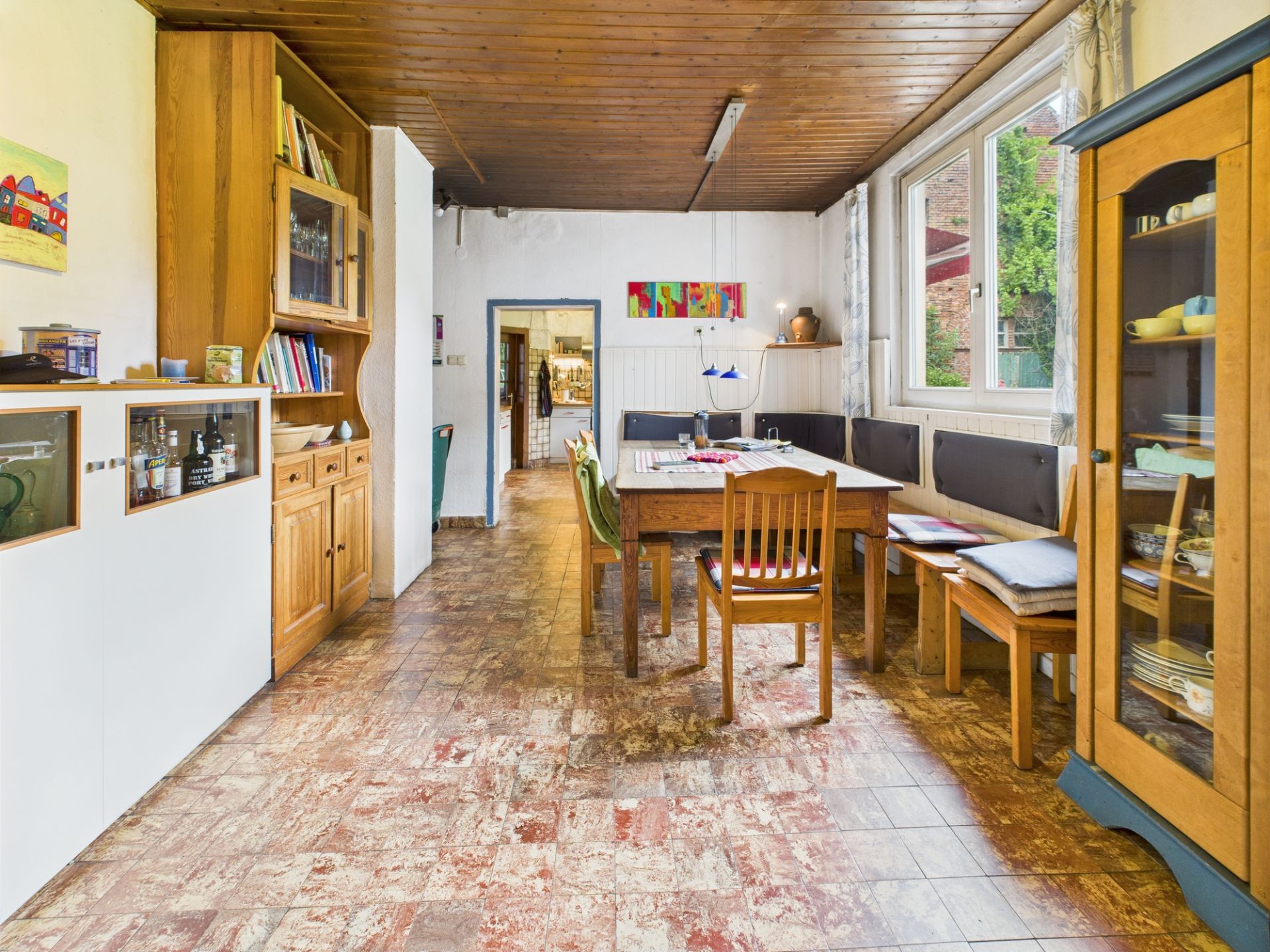 Esszimmer mit Holzmöbeln, großem Tisch, Stühlen, Eckbank und Glasvitrine an einem Fenster.