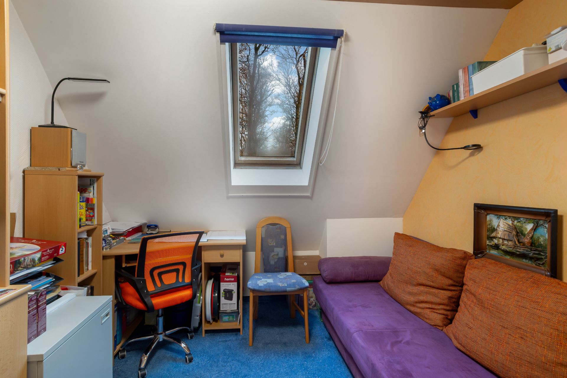 Kleines Zimmer mit Schreibtisch, Stuhl, Regal, lila Sofa und Dachfenster.