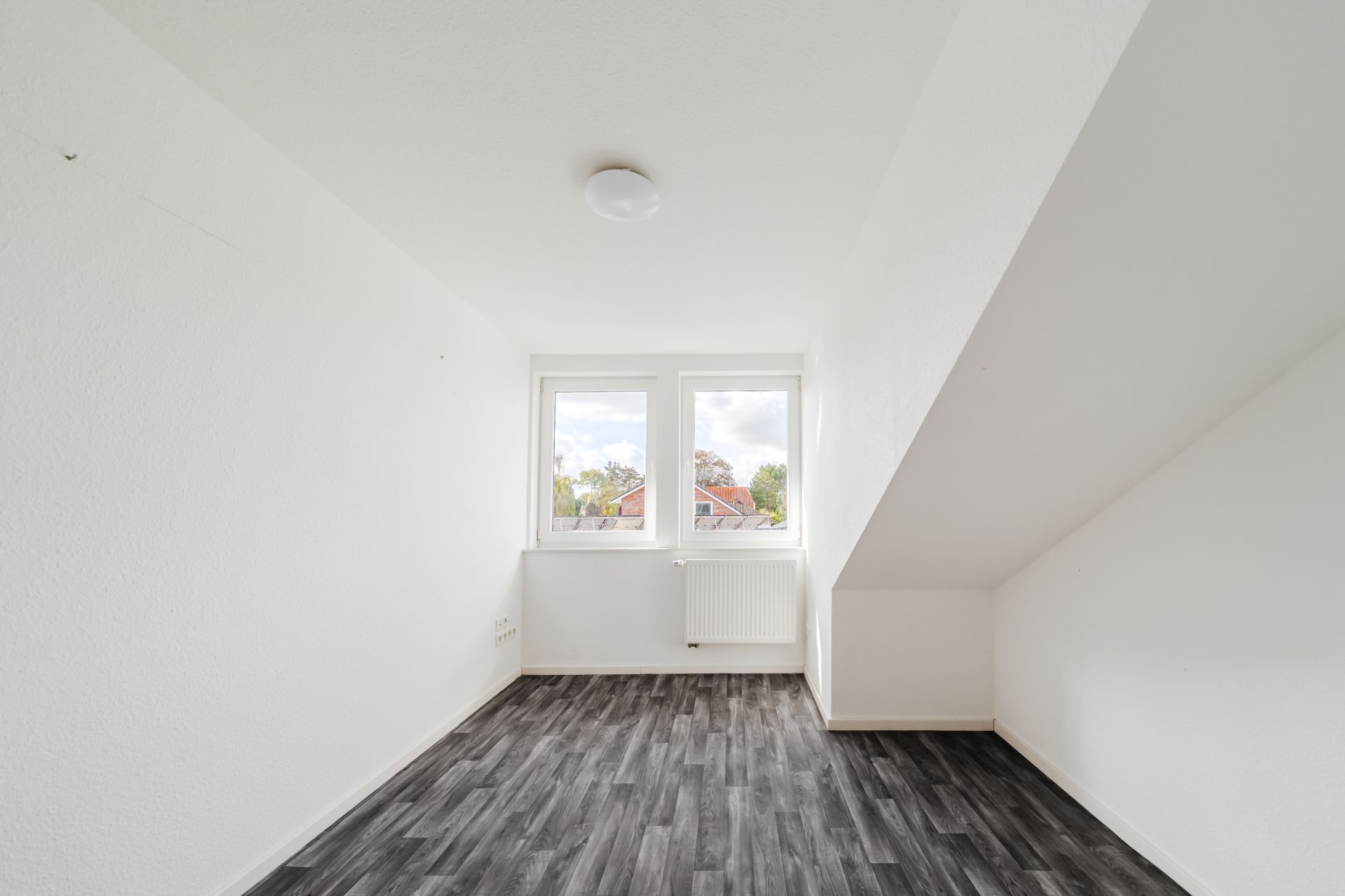 Leeres Zimmer mit zwei Fenstern, weiß gestrichenen Wänden und dunklem Boden.