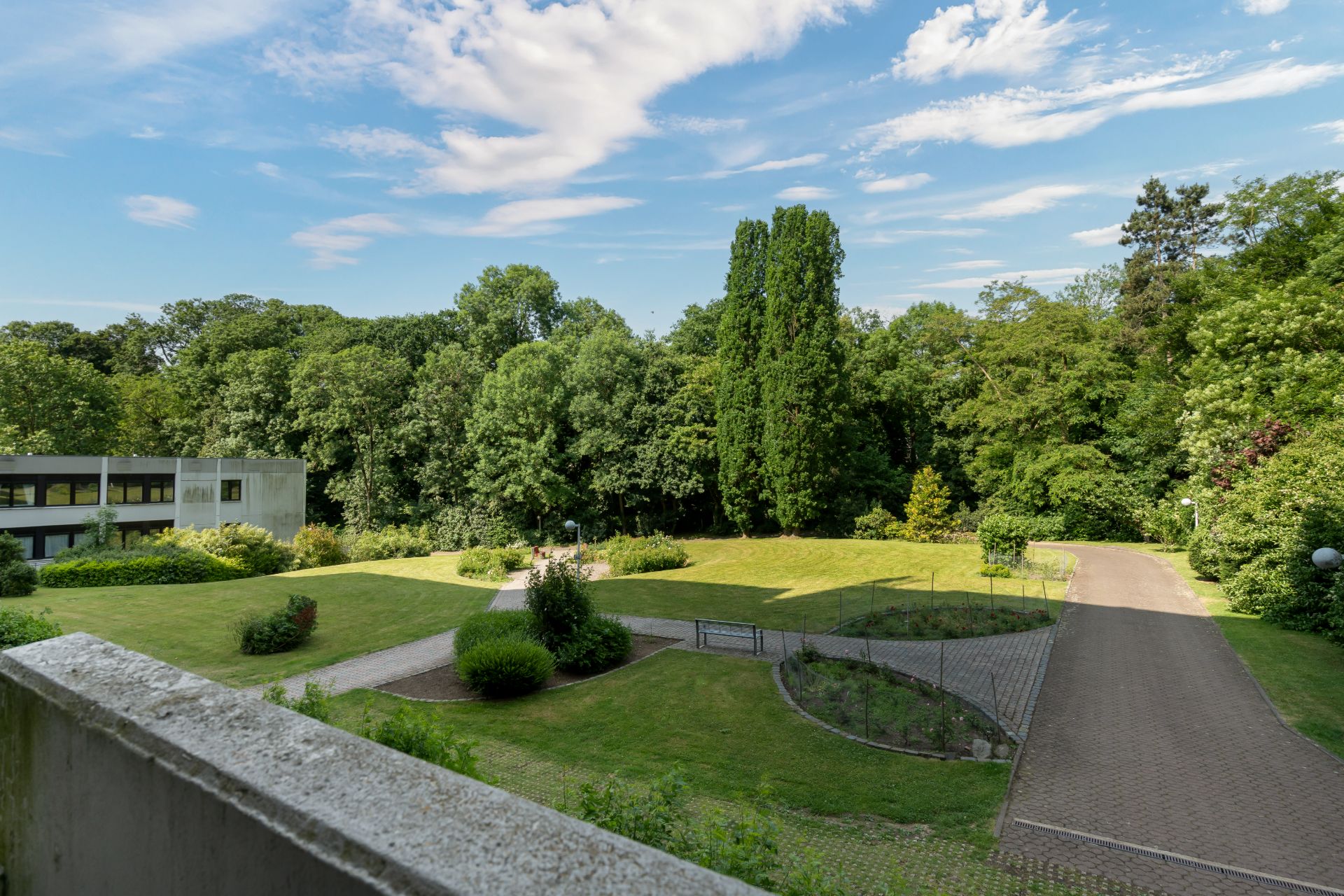 Blick vom Balkon auf einen gepflegten Garten mit Wegen, Bäumen und Rasenflächen.