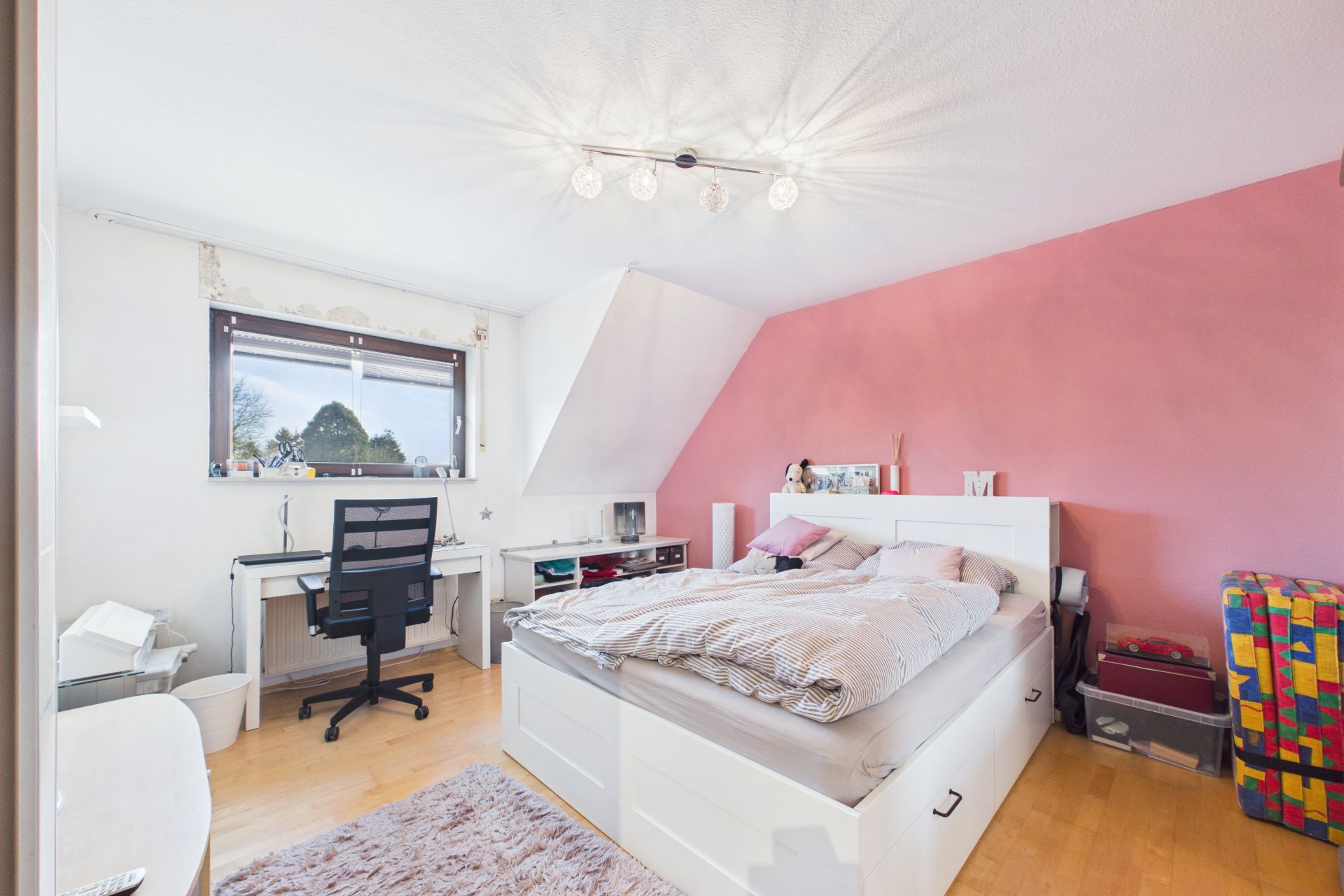 Helles Schlafzimmer mit Doppelbett, Schreibtisch am Fenster, rosa Wand und Holzfußboden.
