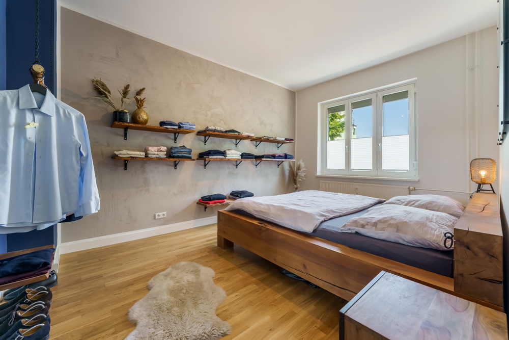 Helles Schlafzimmer mit Holzboden, Doppelbett und Wandregalen mit Kleidung.