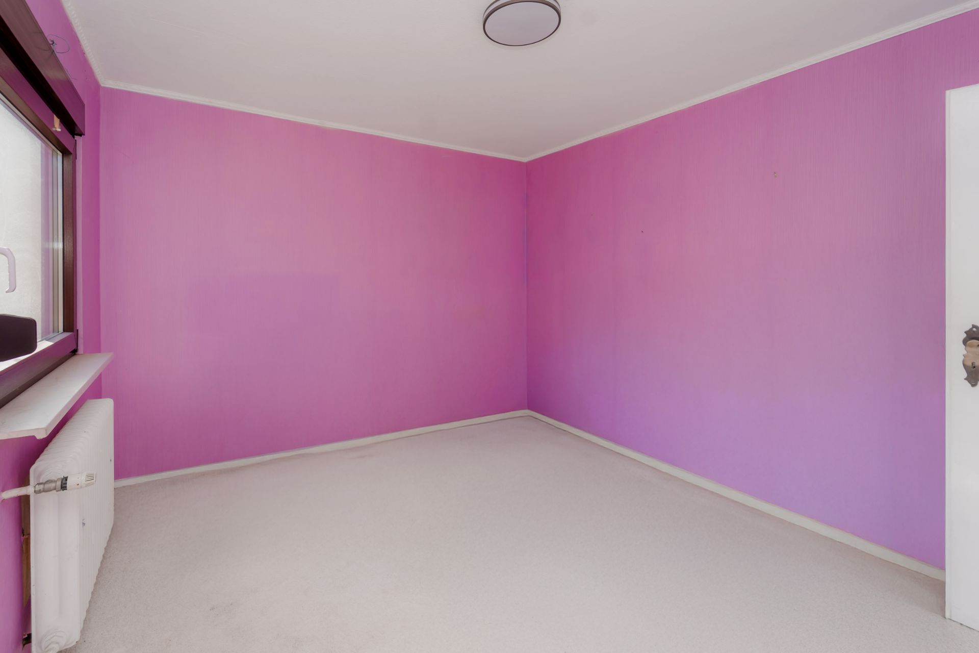 Leerer Raum mit rosa Wänden, weißem Boden, Fenster und Heizung links.