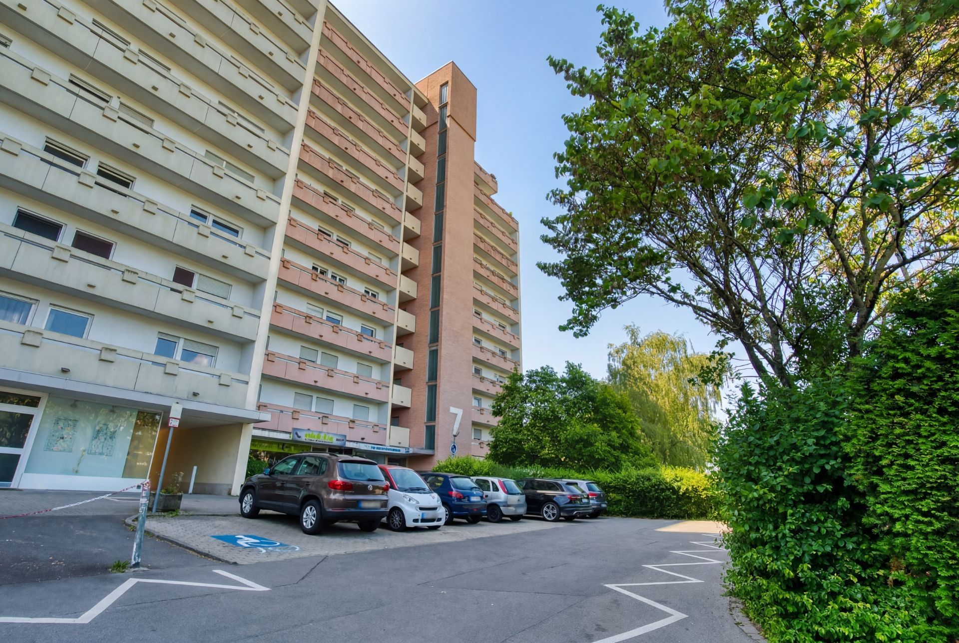 Mehrstöckiges Wohnhaus mit Balkonen und geparkten Autos vor dem Gebäude, daneben Bäume.