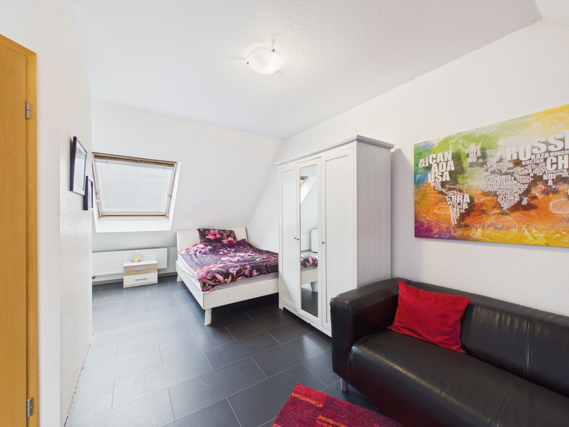 Helles Schlafzimmer mit Bett, Kleiderschrank, schwarzem Sofa und farbigem Weltkartenbild an der Wand.