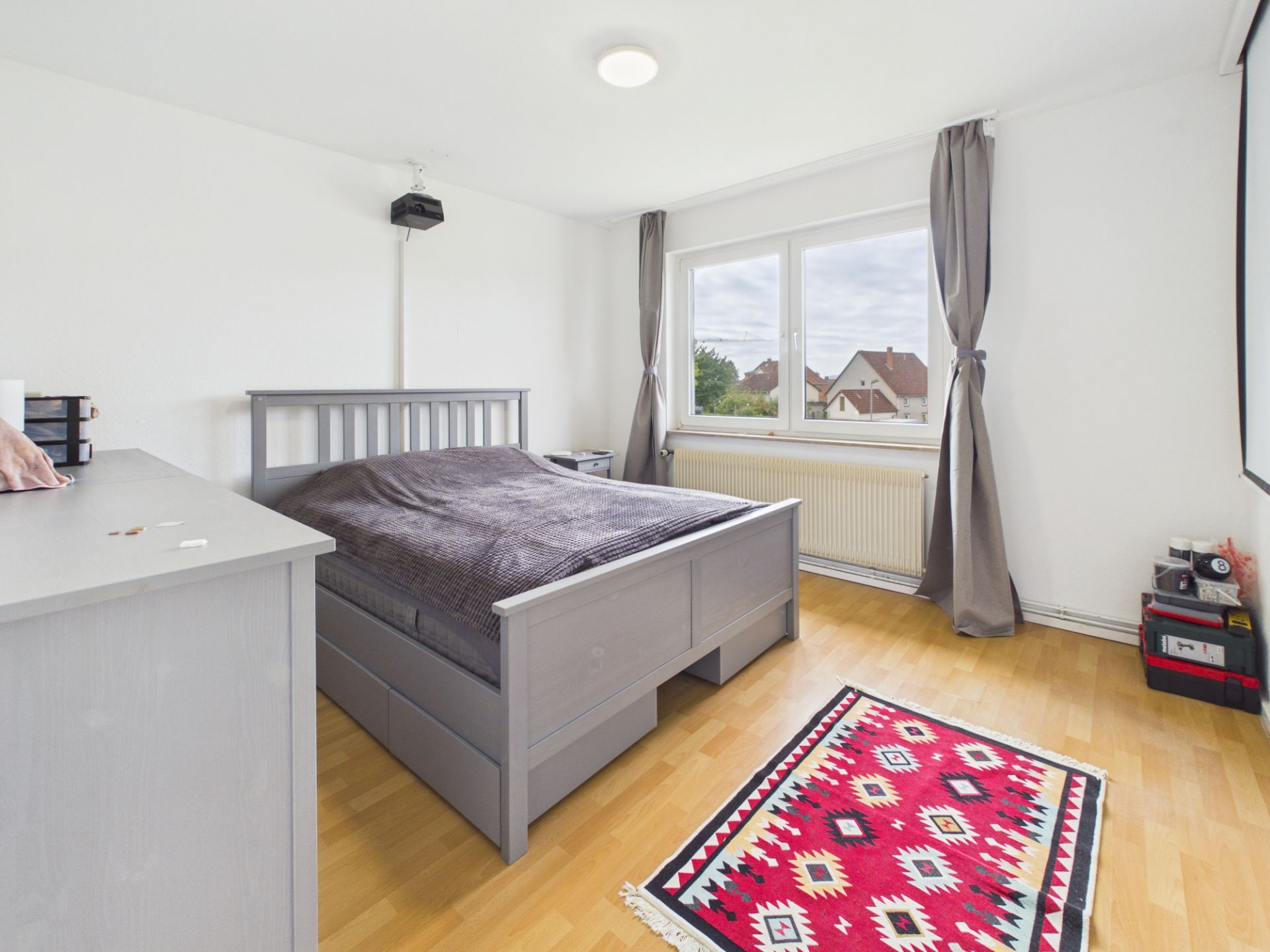 Helles Schlafzimmer mit grauem Bett, Kommode, Teppich und großem Fenster mit Blick nach draußen.