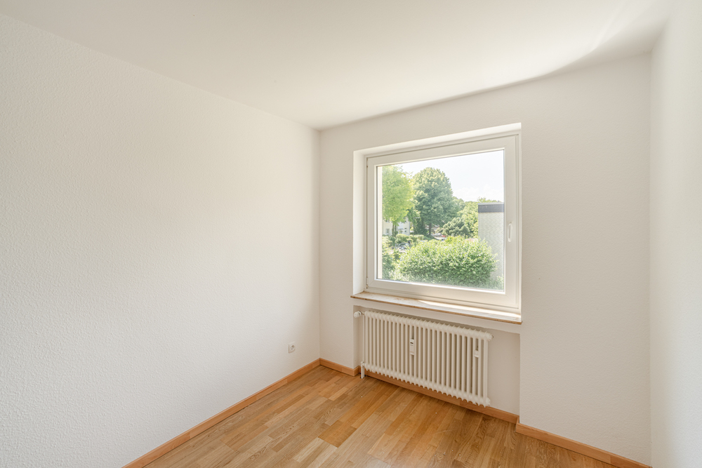 Helles, leeres Zimmer mit Holzfußboden, Fenster und Heizkörper darunter.