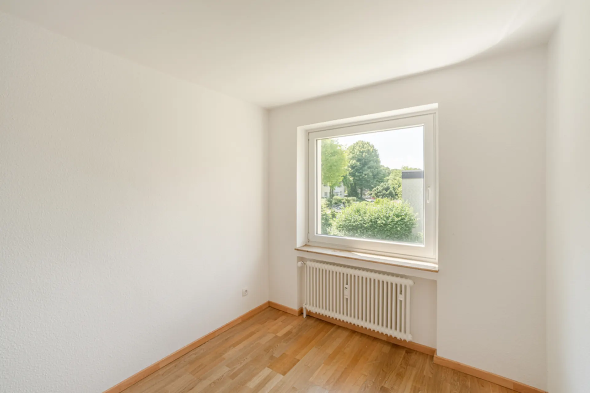 Helles, leeres Zimmer mit Holzfußboden, Fenster und Heizkörper darunter.