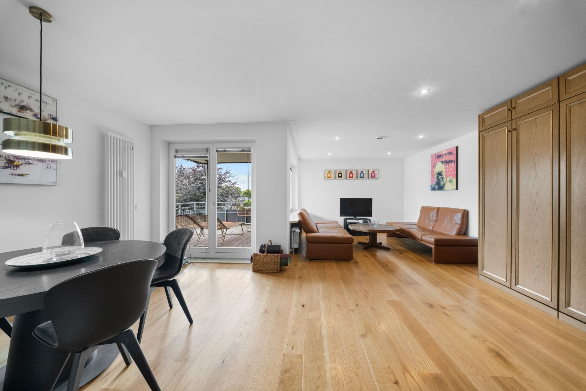 Helles Wohnzimmer mit Holzboden, Essbereich, Sofa, Fernseher und Zugang zum Balkon.