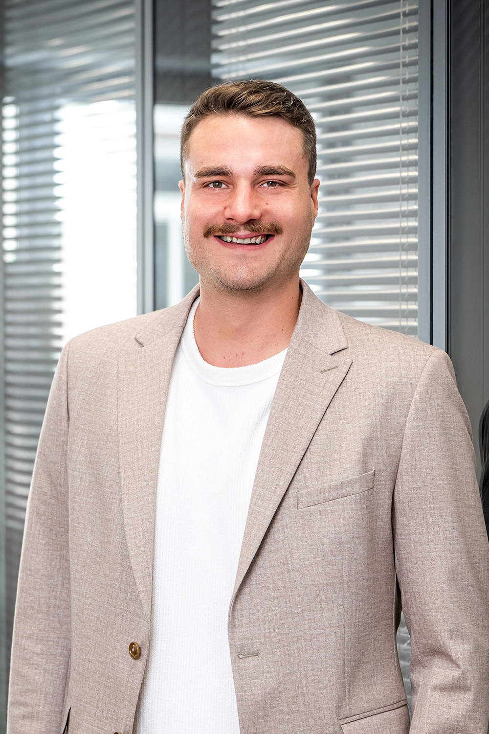 Ein Mann mit hellem Sakko steht vor einer Fensterfront mit Jalousien.