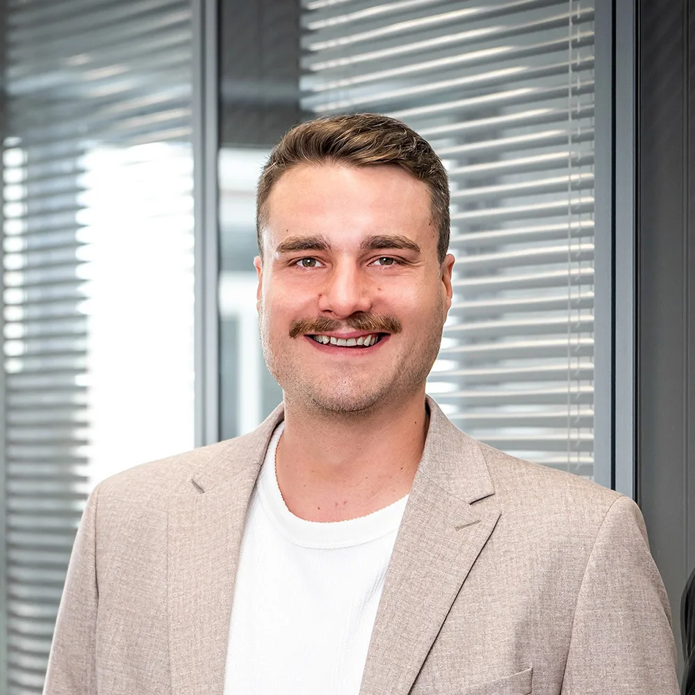 Ein Mann mit hellem Sakko steht vor einer Fensterfront mit Jalousien.