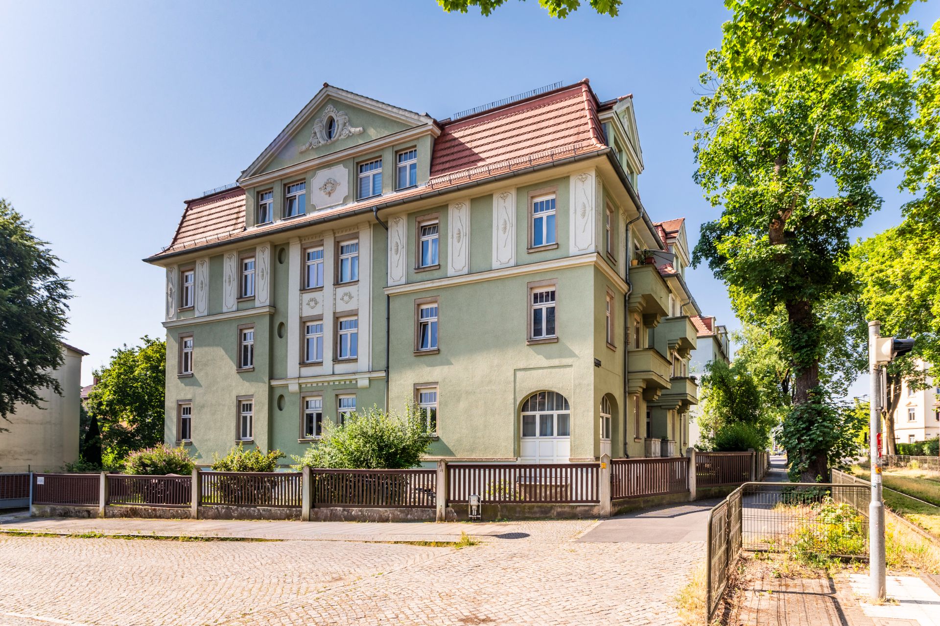 Mehrstöckiges, grünes Wohngebäude mit rotem Dach und verzierten Fassadendetails, umgeben von Bäumen.