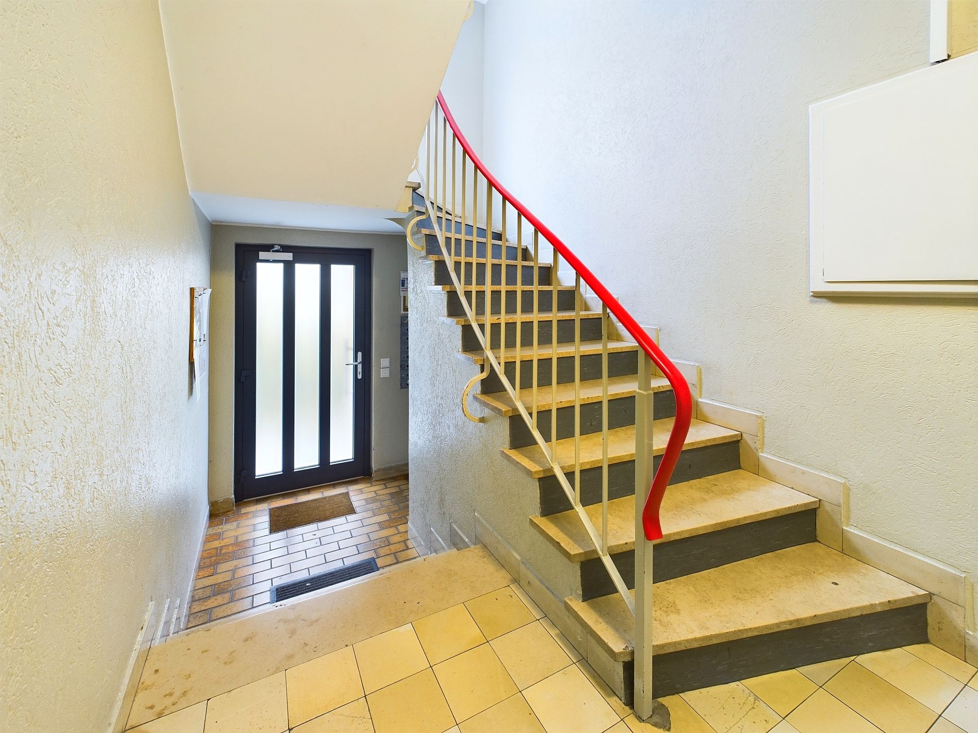 Treppenhaus mit rotem Handlauf und Glastür am Eingang.