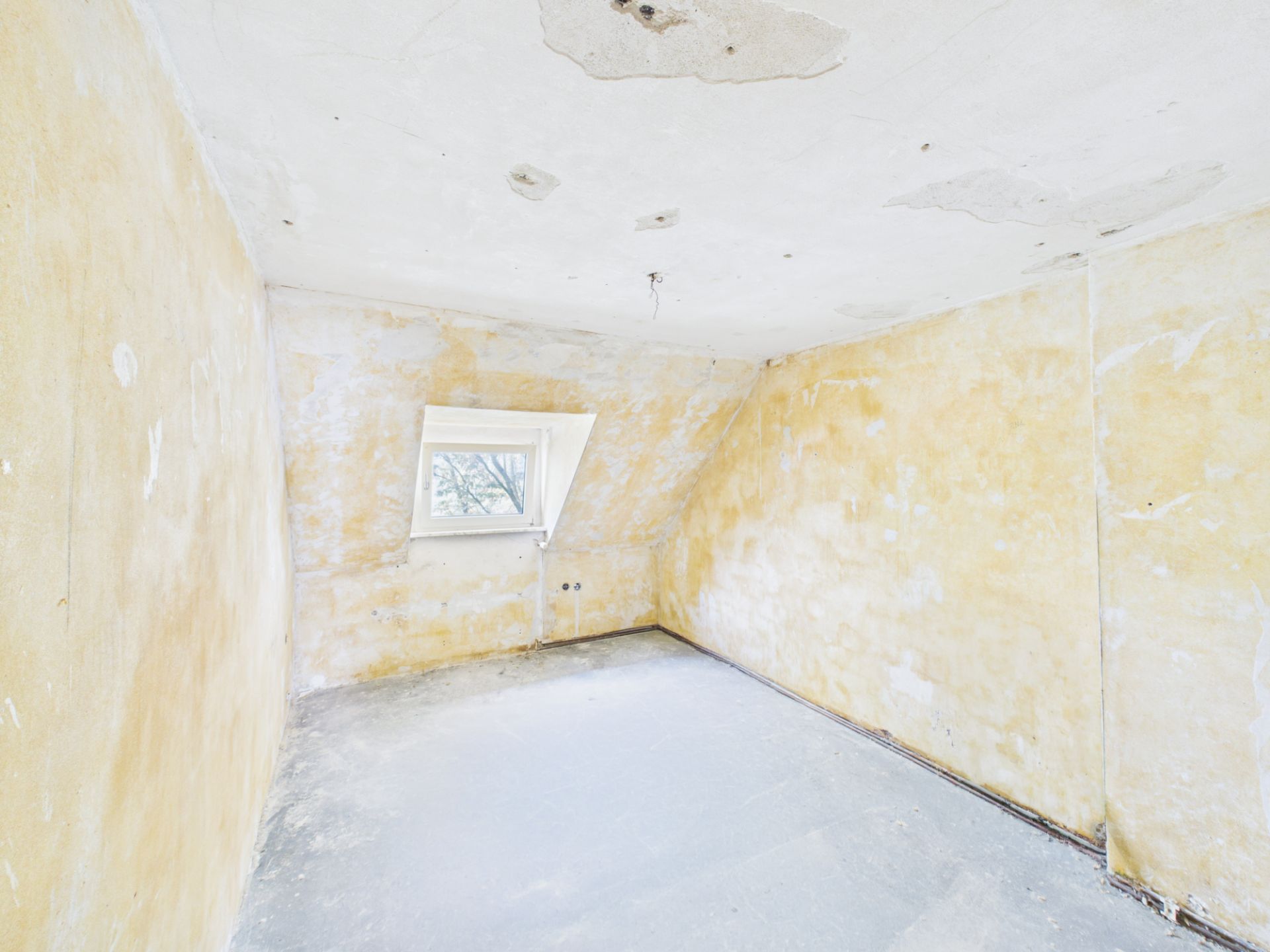 Leerer Raum mit schräger Decke, gelblichen Wänden und einem kleinen Fenster.