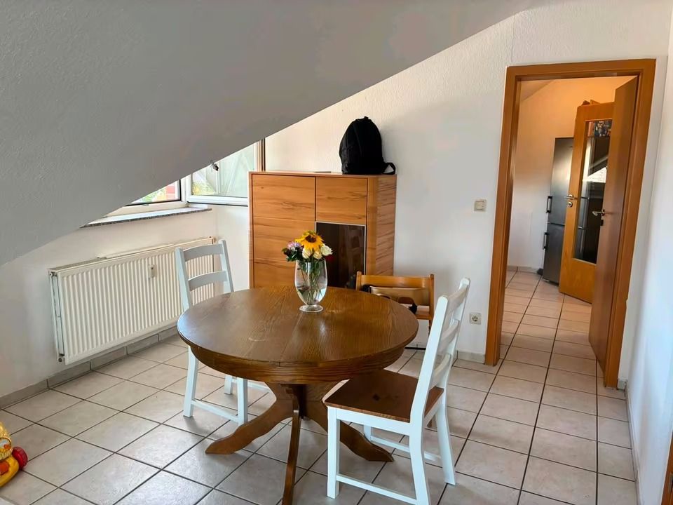 Essbereich mit rundem Holztisch, drei Stühlen, Kommode und Vase mit Blumen, unter Dachschräge.
