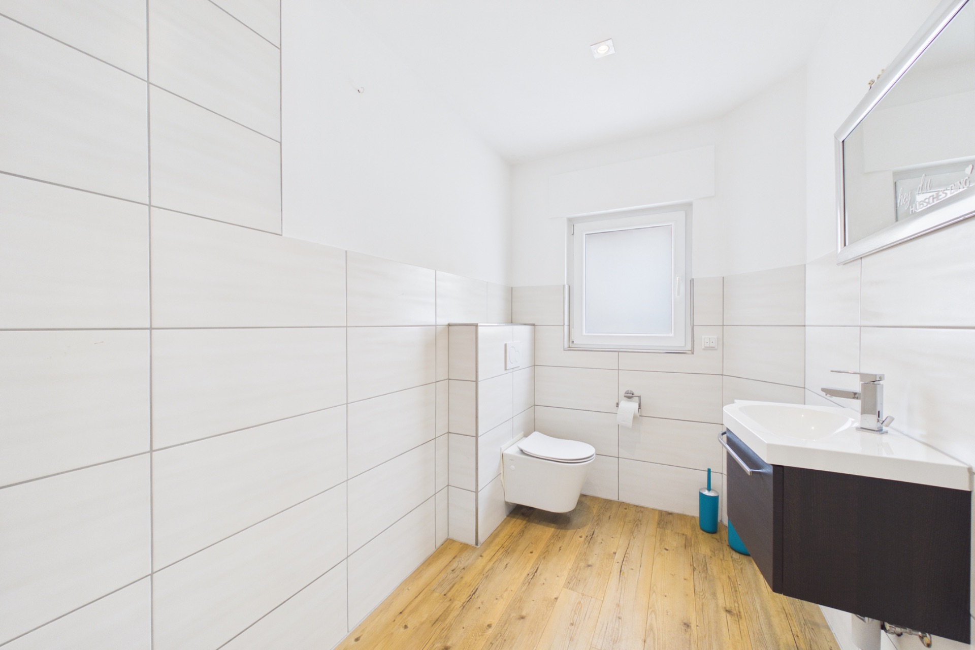 Modernes Badezimmer mit Wandtoilette, Waschbecken, Spiegel und Fenster, heller Fliesenwand und Holzfußboden.