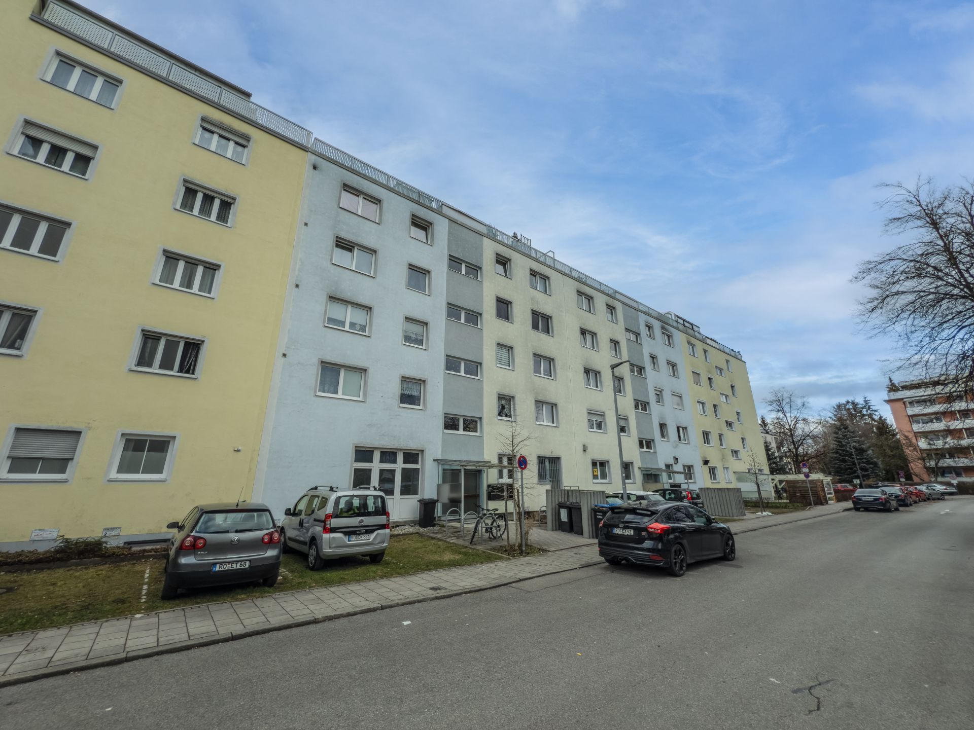 Mehrgeschossiges Wohnhaus mit Autos und Fahrrädern davor an einer ruhigen Straße.