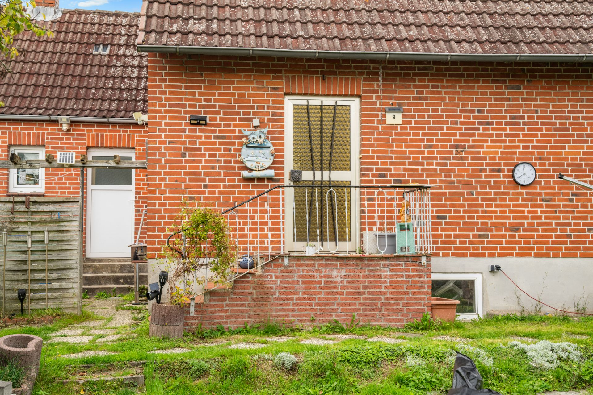Backstein-Hauseingang mit kleinem Treppenaufgang, Geländer und Garten im Vordergrund.