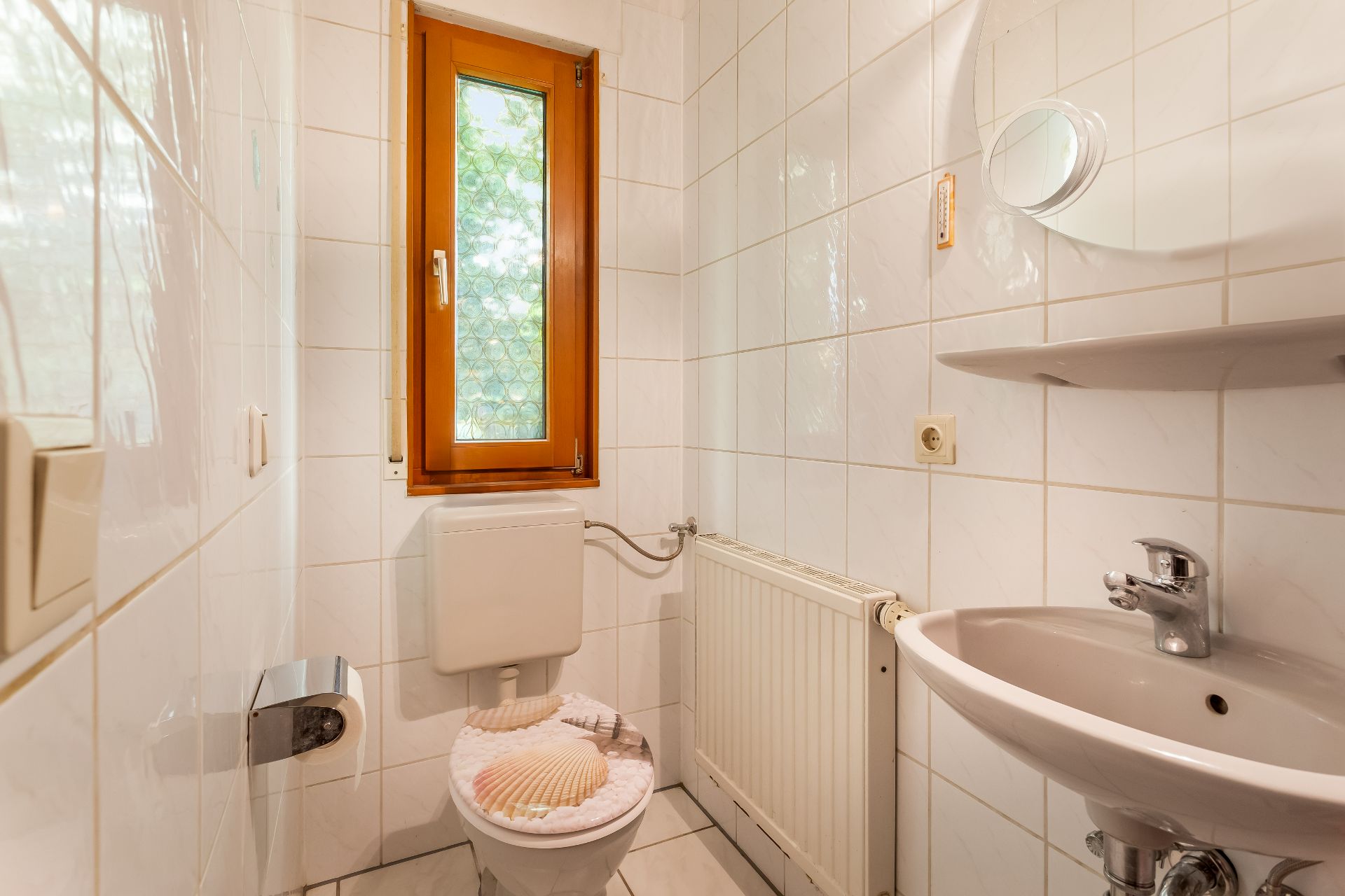 Kleines Badezimmer mit Toilette, Waschbecken, Fenster und Heizung. Muscheln auf dem Toilettendeckel.