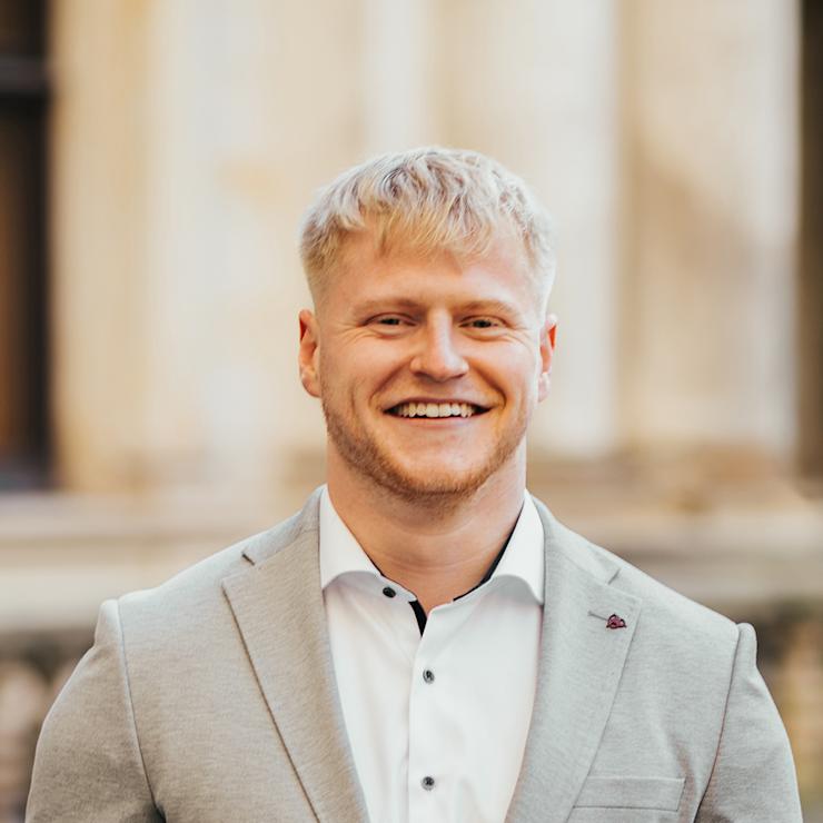 Ein junger Mann mit blondem Haar lacht und trägt einen grauen Anzug mit weißem Hemd.