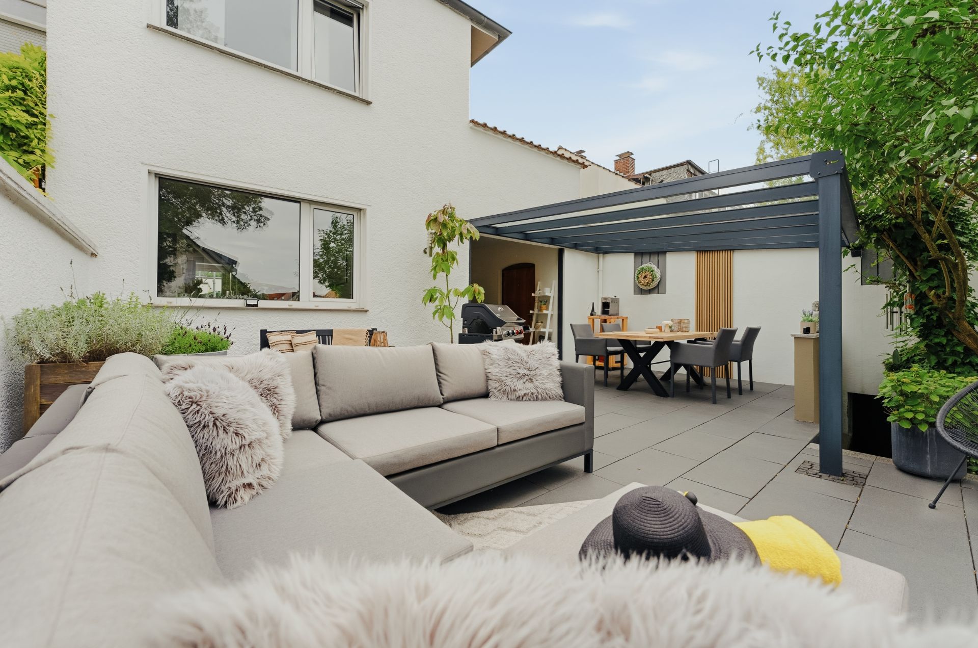 Moderne Terrasse mit Sitzgruppe, Esstisch unter Pergola und Pflanzen an einem Wohnhaus.