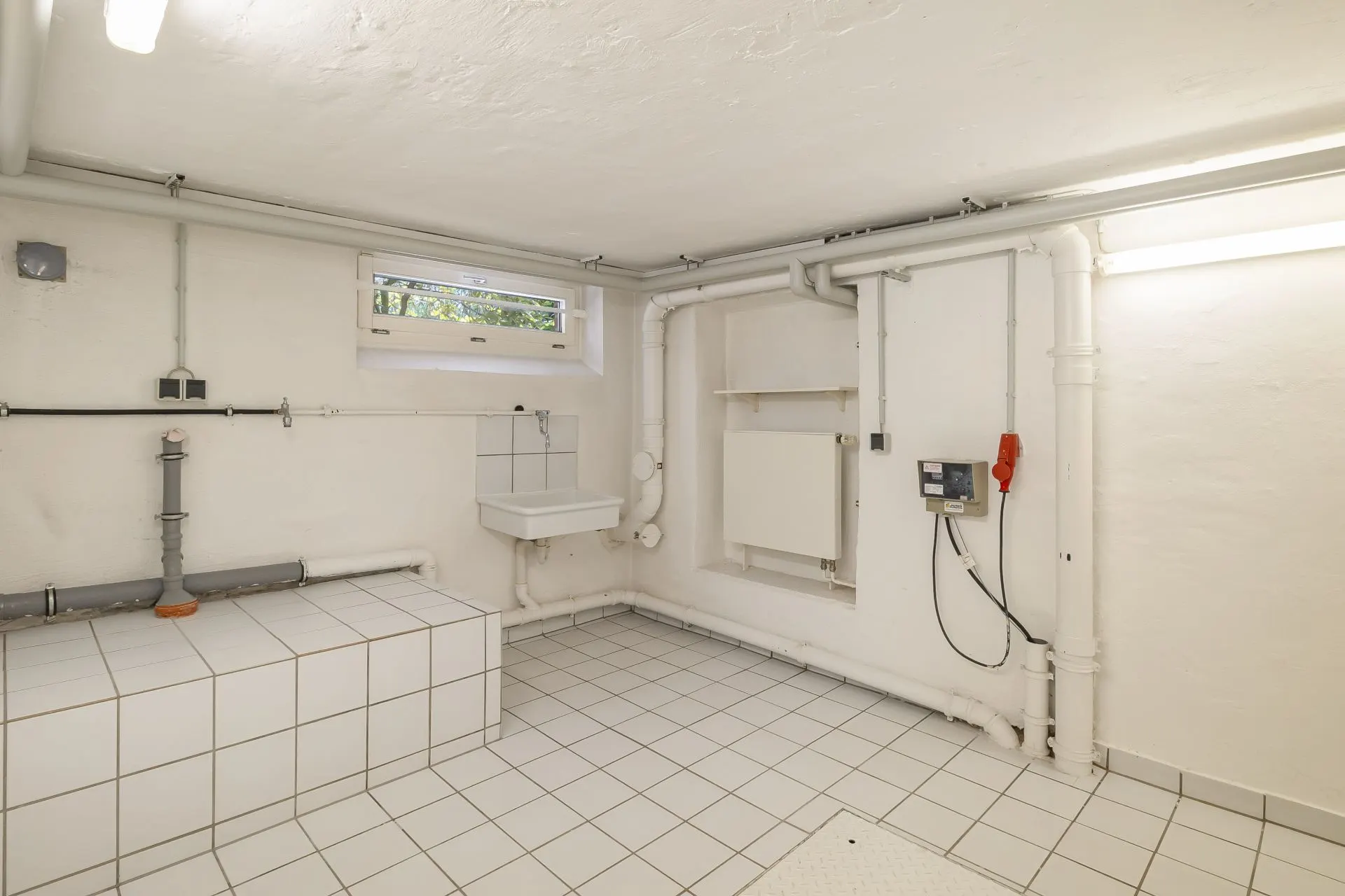 Heller Kellerraum mit Fliesenboden, Waschbecken, Rohren und kleinem Fenster.