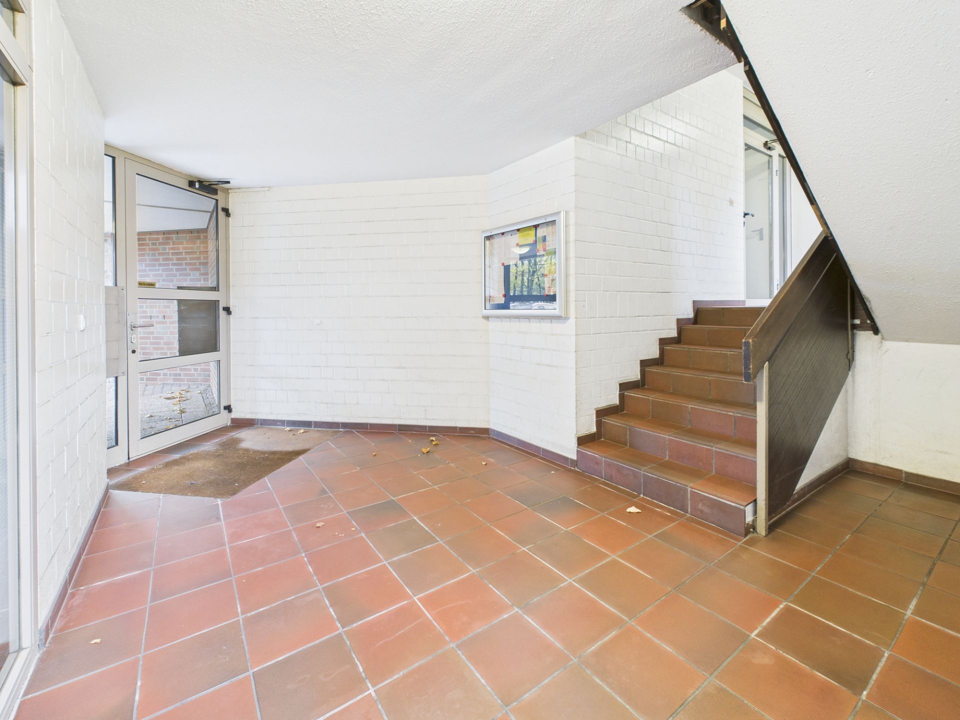 Eingangsbereich mit braunen Fliesen, Treppe und Glastür, weiße Wände.
