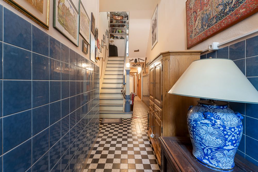 Flur mit Schachbrettboden, blauen Fliesen, Holzschrank, Tischlampe und Treppe nach oben.