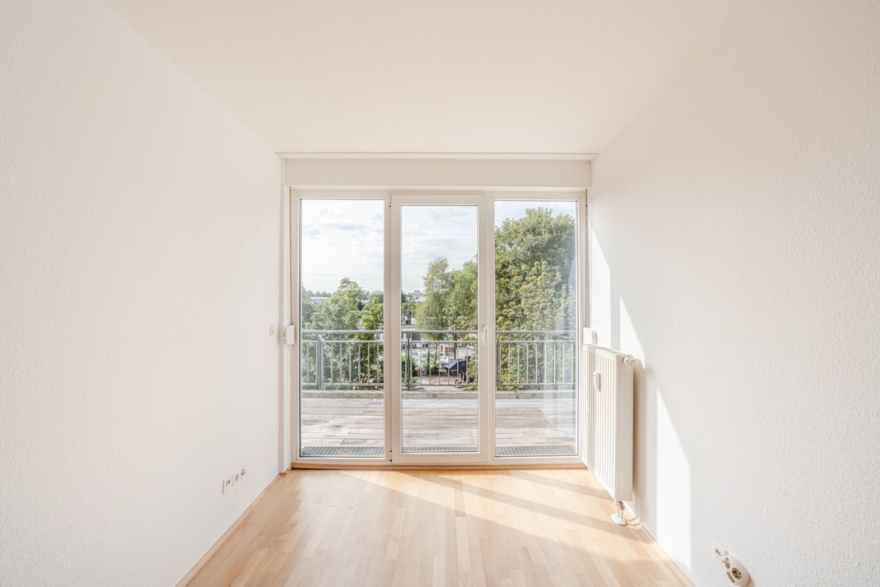 Heller Raum mit Parkettboden und großer Glas­tür zu einem Balkon mit Blick auf Bäume.