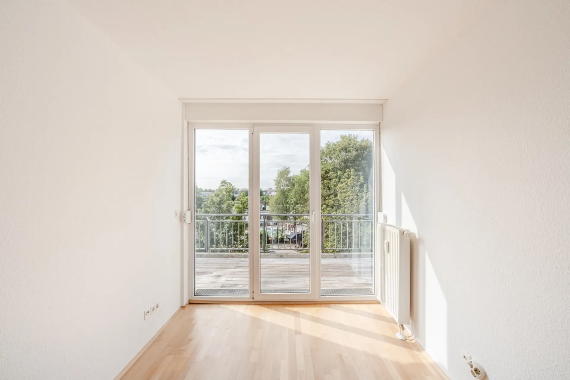 Heller Raum mit Parkettboden und großer Glastür zu einem Balkon mit Blick auf Bäume.