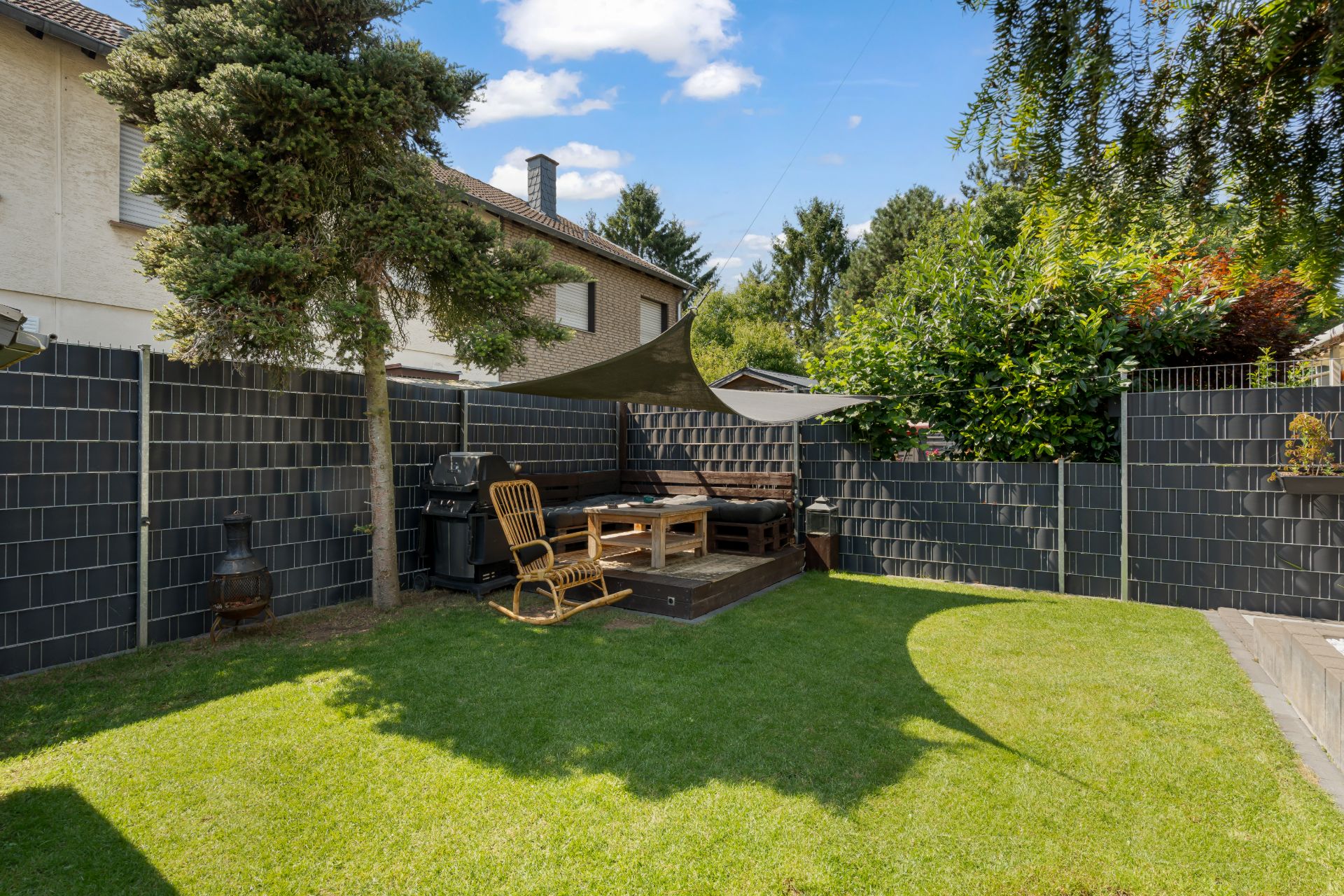 Garten mit Rasenfläche, Sitzbereich auf Holzterrasse, Sonnensegel und umlaufendem Zaun.