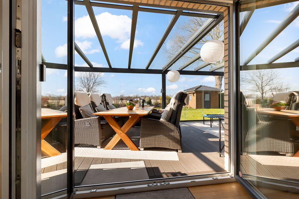 Blick durch offene Glastür auf überdachte Terrasse mit Sitzgruppe und Garten.