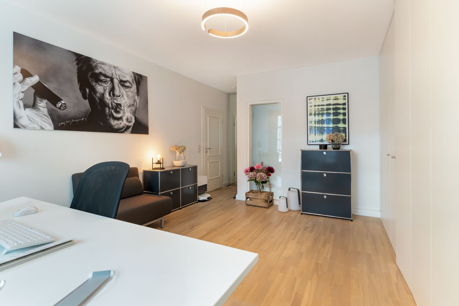 Helles Arbeitszimmer mit Schreibtisch, Stuhl, Kommoden und Wandbildern auf Holzfußboden.